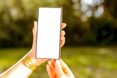 Beyaz ekranlı akıllı telefon, arka planda doğal bahçedeki kadınların ellerinde. Doğal güneş ve gün ışığı. Boşluğu kopyala.