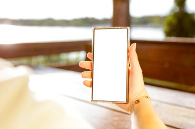 Elinde fotokopi alanı olan beyaz ekranlı telefonu tutan kadın. Arka planda göl ve gökyüzü bulanık. Uzak çalışma yaşam tarzı ve doğa konseptinde yaratıcı düşünce.