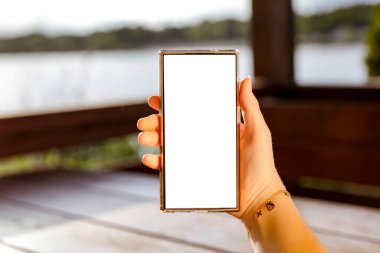 Elinde fotokopi alanı olan beyaz ekranlı telefonu tutan kadın. Arka planda göl ve gökyüzü bulanık. Uzak çalışma yaşam tarzı ve doğa konseptinde yaratıcı düşünce.