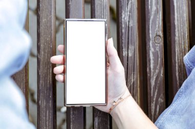 Akıllı telefon, genç bir bayanın elinde güneşli bir parktaki ahşap bankta oturuyor. Modern teknolojiyle doğal çevreyi harmanlayan kavramsal bir görüntü. Metin için alanı kopyala.