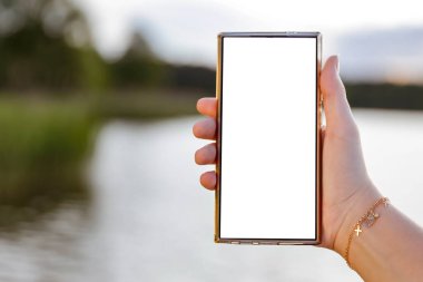 Elinde fotokopi alanı olan beyaz ekranlı telefonu tutan kadın. Arka planda göl ve gökyüzü bulanık. Uzak çalışma yaşam tarzı ve doğa konseptinde yaratıcı düşünce.