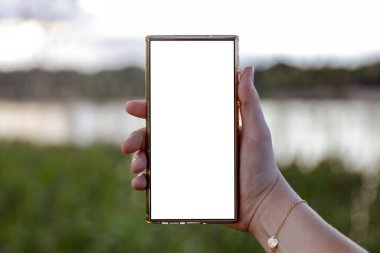 Elinde fotokopi alanı olan beyaz ekranlı telefonu tutan kadın. Arka planda göl ve gökyüzü bulanık. Uzak çalışma yaşam tarzı ve doğa konseptinde yaratıcı düşünce.