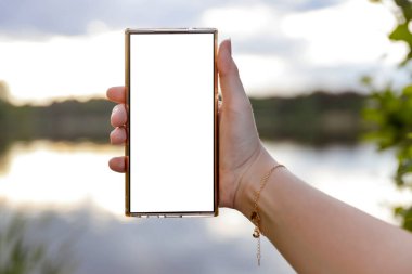 Elinde fotokopi alanı olan beyaz ekranlı telefonu tutan kadın. Arka planda göl ve gökyüzü bulanık. Uzak çalışma yaşam tarzı ve doğa konseptinde yaratıcı düşünce.