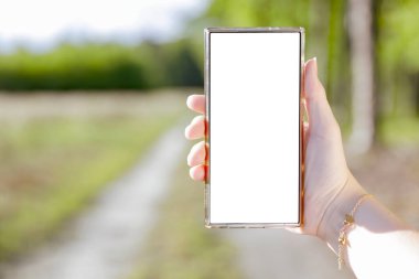 Güneşli bir orman arka planına sahip akıllı bir telefonu tutan bir kadının yakın çekimi. Doğal çevreye sahip modern teknoloji, uzak çalışmayı sembolize ediyor, açık havada yaratıcı düşünme..