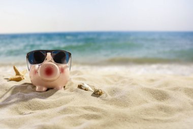 Kumsalda güneş gözlüklü domuz. Domuz kumbarası, bulanık plaj, deniz ve gökyüzü arka planı, ürün yerleştirme ve metin için alan. İş ve tatil konsepti için para biriktirme.