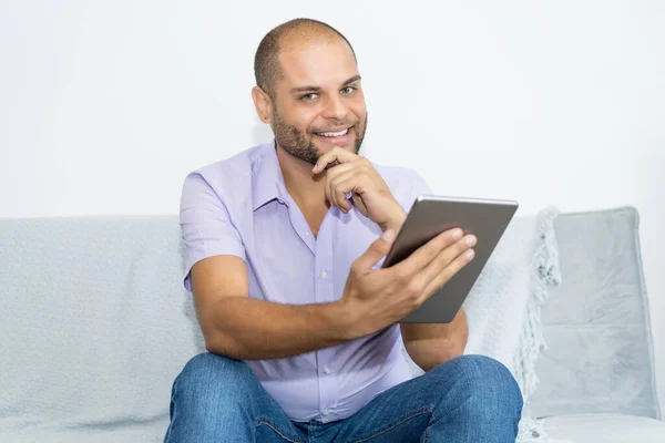 Laughing man with bald and beard working at digital tablet indoors at home at couch
