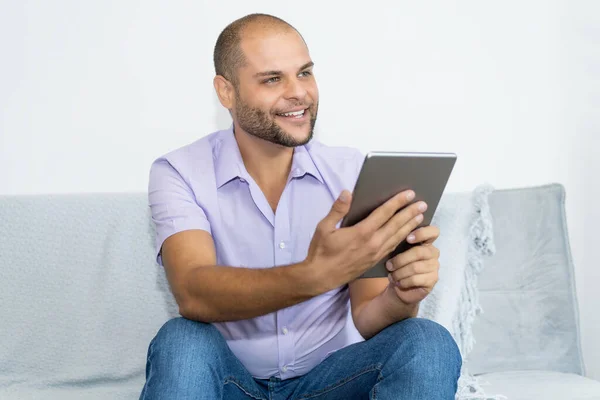 Man with bald and beard working at digital tablet indoors at home at couch