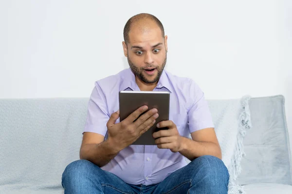 Man with bald and beard watching movie on digital tablet indoors at home at couch