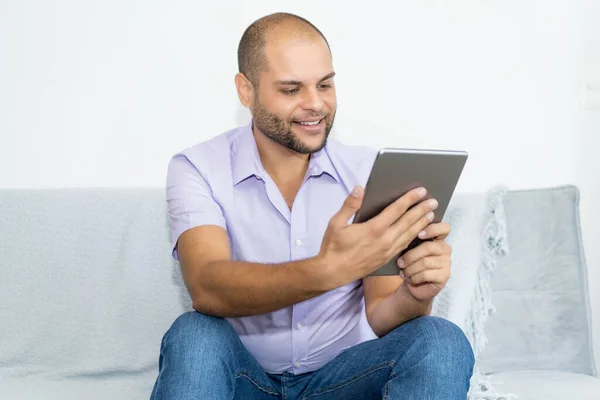 Happy man with bald and beard reading news on digital tablet indoors at home at couch