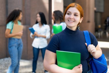 Laughing german female student with red hair and group of young adults in front of university