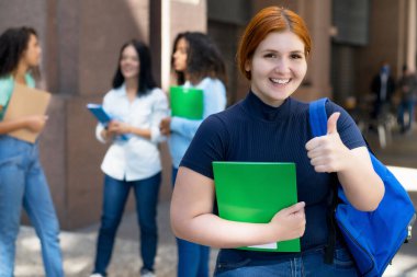 Successful german female student with red hair and group of young adults in front of university