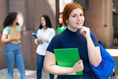 Beautiful german female student with red hair and group of young adults in front of university