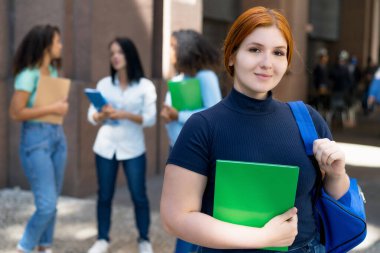 Young german female student with red hair and group of young adults in front of university
