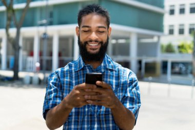 Handsome black man with beard  laughing and sending message to girlfriend outdoor in summer in city