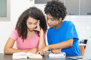 Focused hispanic and african american female student preparing for graduation indoors at desk at home