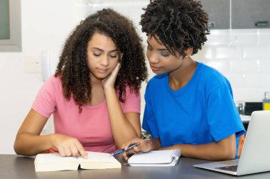 Smart hispanic and african american female student preparing for graduation indoors at desk at home