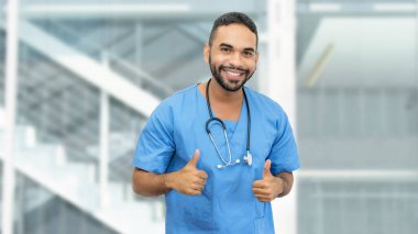 Sakallı ve steteskoplu Meksikalı erkek hemşire ya da doktor klinikte baş parmak gösteriyor.