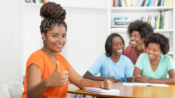 大学の教室でアフリカ系アメリカ人の学生を学ぶグループと親指を示すデスクで成功した黒人女性学生 — ストック写真