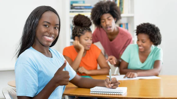 在课桌前展示大拇指的漂亮黑人女生与一群在大学课堂上学习的非洲裔美国学生 — 图库照片
