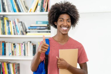 Sırt çantası ve evrakları olan zeki Afro-Amerikan erkek öğrenci üniversite sınıfında baş parmağını gösteriyor.