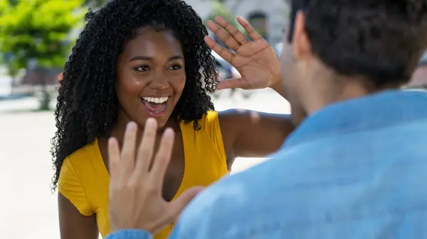 Happy black female young adult giving high five to latin american friend outdoor in summer in city