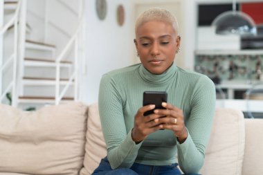 Burun piercingli, siyah saçlı, evde evdeki kanepede telefonla mesaj okuyan Afro-Amerikalı bir kadın.
