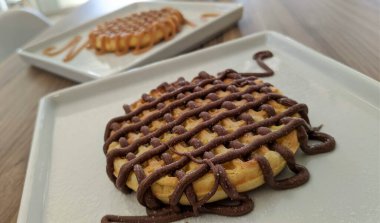 Beyaz kare tabakta çikolatalı waffle ve arka planda karamel kaplı waffle. 
