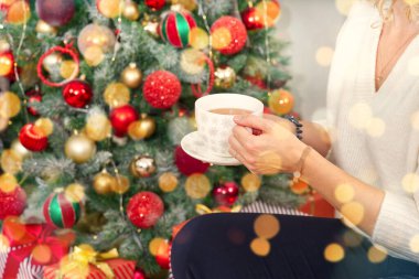 Beyaz kazaklı bir kız elinde bir fincan sıcak kahve ya da bir Noel ağacının arka planında çay tutuyor. Sıcak Noel atmosferi. Noel için alkolsüz sıcak bir içki. Yaşam biçimi