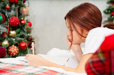 Mutlu genç kız elinde kalem ve defterle dilek listesi hazırlıyor ya da yılbaşı ağacı üzerinde yeni yıl için yatakta liste yapıyor. Genç kız Noel zamanı rüya görüyor. xmas tatil konsepti
