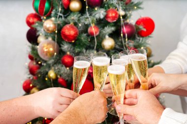 Yeni yıl ya da Noel kutlaması. Şampanya kadehlerini tutan eller. Arka planda çelenkler olan Noel ağacı.