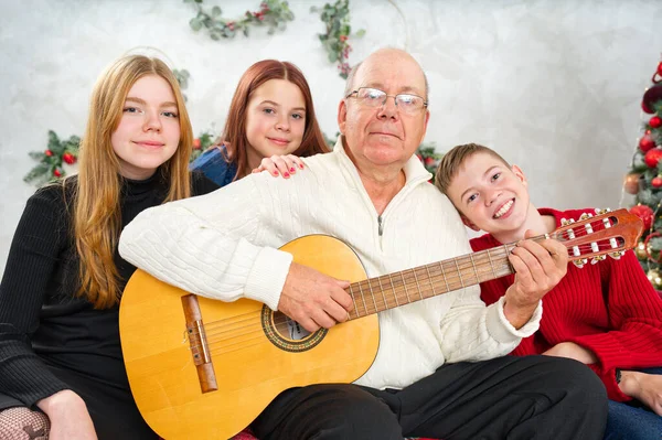 Grandfather playing guitar during christmas for grandchildren. Happy man 60s playing guitar christmas songs. Happy christmas atmosphere.