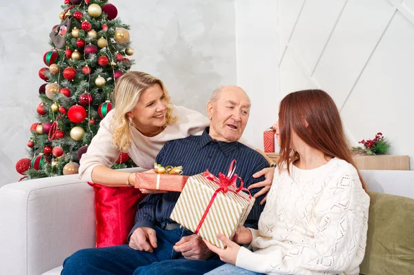 Granddaughters give a gift to grandfather. Happy senior man embracing his granddaughter while receiving a gift on Christmas at home. Happy family grandfather with cute excited granddaughter.