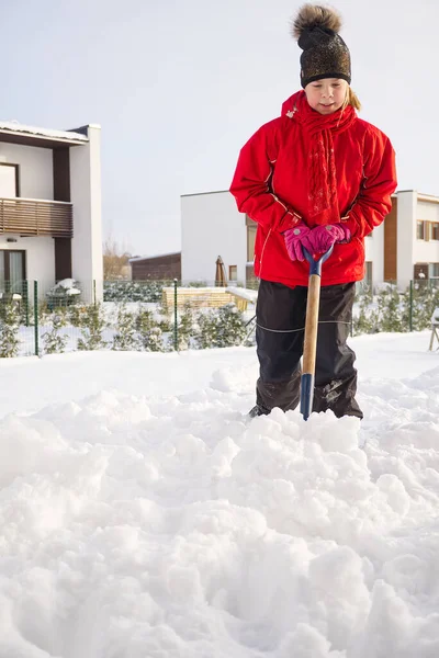 Girl shoveling snow on home drive way. Beautiful snowy garden or front yard. Teenager removing snow with a shovel in the winter. Little Girl in red jumpsuit cleans snow big shovel. Snow removal after