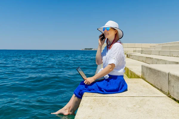Work from anywhere. Side view of young woman, female freelancer in straw hat working on laptop while sitting on the beach. young woman in sitting on stone at sea and remotely working on laptop