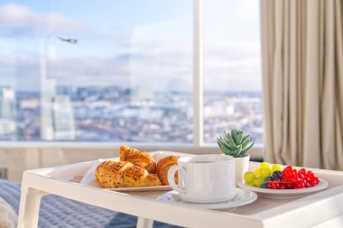 White Tray with tasty breakfast on bed in light bedroom with big windows in skyscraper. white table with tasty breakfast in bed at bed room in skyscraper