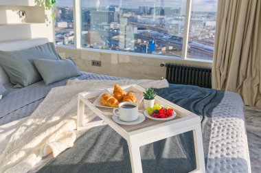 White Tray with tasty breakfast on bed in light bedroom with big windows in skyscraper. white table with tasty breakfast in bed at bed room in skyscraper