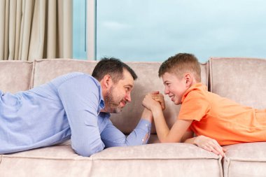 Handsome young father and his cute son competing in arm wrestling while lying on a sofa. Father and son quality time