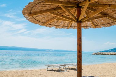 Güneşli bir günde deniz kıyısındaki altın kumsalda saman plajı şemsiyesi ile çekilen sakin bir plaj sahnesi. Açık mavi gökyüzü ve sakin sular mükemmel bir sahil kaçışı için sakin bir atmosfer yaratır..
