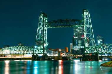 Queens Köprüsü olarak da bilinen Koninginnebrug Köprüsü 'nün, Rotterdam' ın dingin sularına yayılan, parlak renklerle aydınlatılmış uzun gece fotoğrafı..