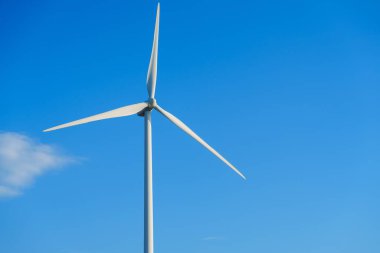 Temiz enerjiyi sembolize eden, açık mavi gökyüzüne karşı üç bıçaklı büyük beyaz bir rüzgar türbini. Sürdürülebilir elektrik üretimi için rüzgar enerjisinden faydalanıyor ve daha yeşil bir geleceğe katkı sağlıyor.