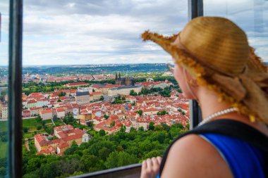Şık bir hasır şapka giyen bir kadın turist Prag 'ın çarpıcı şehir manzarasına Prag Kalesi' nin ikonik kulesinin penceresinden bakıyor. Manzara şehrin zengin tarihini ve güzelliğini gözler önüne seriyor..