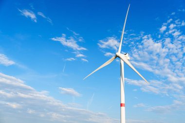 Uzun, beyaz bir yel değirmeni açık mavi bir gökyüzüne karşı duruyor. Bıçakları hızla dönüyor ve yenilenebilir enerjinin güzelliğini gösteriyor. Düşük açı, yel değirmenlerinin yüksekliğini ve izolasyonunu arttırır..