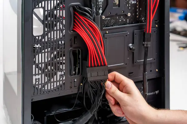 stock image A hand connects a black ATX power supply cable to an Asus motherboard inside a sleek black case. The motherboard features red accents on a black PCB, with a tempered glass side panel for visibility.