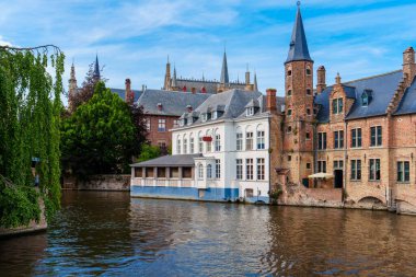 Bruges, Belçika 'daki bir kanal manzarası. Sakin su kıyılar boyunca büyüleyici binaları yansıtır, berrak mavi bir gökyüzü ve yumuşak bulutlar dingin atmosferi güçlendirir..