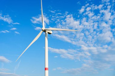 Yalnız bir rüzgar türbini, kabarık bulutlu mavi bir gökyüzüne karşı dimdik ayakta duruyor. Türbinler, rüzgarın enerjisini yakalayıp temiz elektriğe dönüştürmek için yavaş döner..