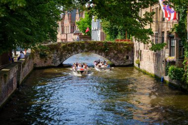 July 2024, Brugge, A scenic canal in Bruges, Belgium features a boat tour under a stone bridge, surrounded by historic buildings and lush foliage. The calm water reflects the vibrant sky, enhancing clipart