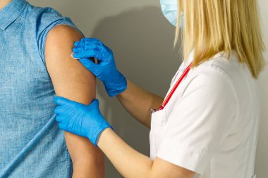 Beyaz önlüklü ve mavi eldivenli genç bir kadın bir adamın koluna iğne yapıyor. Adam mavi bir gömlek giyiyor ve arka plan bulanık. Kadın maske takıyor..