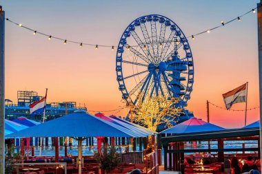 Güneşin batışına karşı aydınlatılmış bir lunaparkta çarpıcı bir dönme dolap, masaları ve şemsiyeleri olan rahat bir restoran ziyaretçileri manzaranın tadını çıkarmaya davet ederken, uzun bir süre ayakta kalır. Lahey