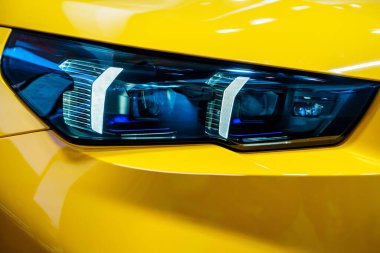 Görüntü sarı bir arabanın farlarının yakın çekimini gösteriyor. Farlar açık ve pırıl pırıl parlıyor. Araba temiz ve cilalı, ve boya güneşte parlıyor..
