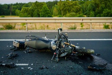 Bir motosiklet yolda hasar görmüş, enkaz ve kazdan arta kalanlarla çevrili. Bölge ağaçlarla kaplı ve güvenlik için bir bariyer var..